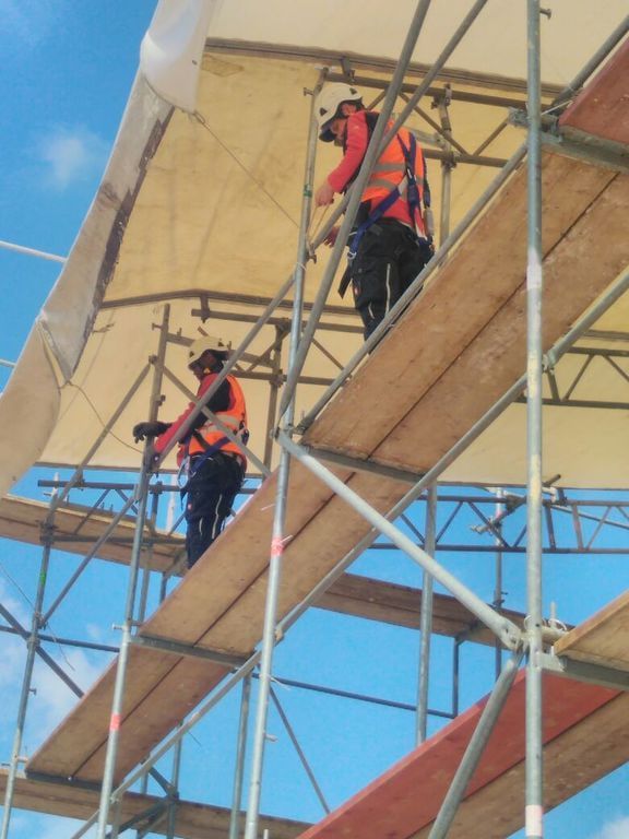 Zwei Gerüstbauer in oranger Warnkleidung mit Helm und Auffanggurt arbeiten auf der obersten Lage eines Fassadengerüsts unter einer Dachkante.